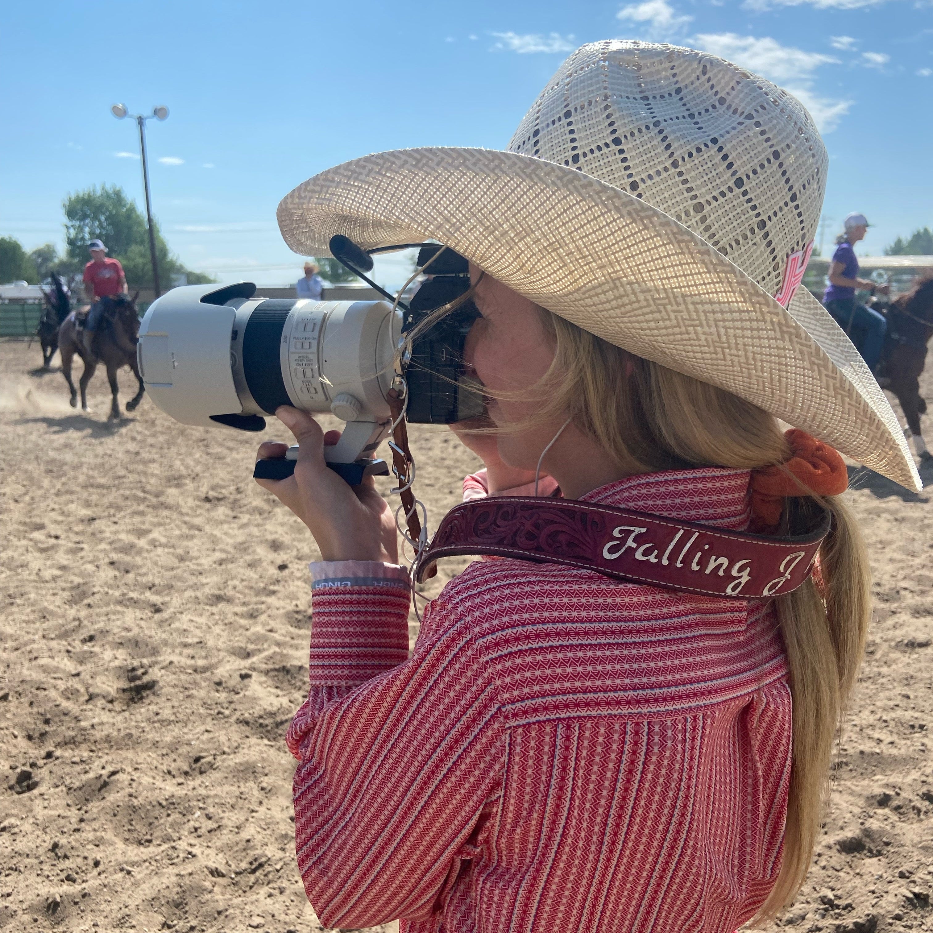 Person in cowboy hat and red checkered shirt holding a camera with a lens, with people on horses in the background.